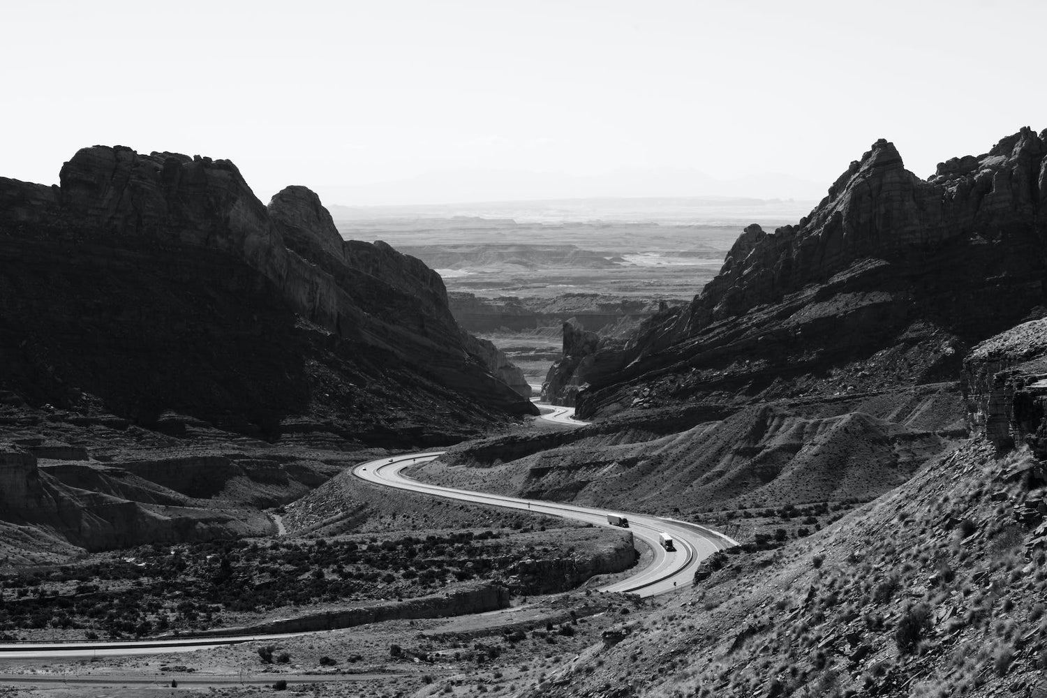 grayscale photo of roadway surrounded with rocky mountains